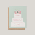 A greeting card with a kraft envelope, featuring a pastel mint background and an illustration of a three-tier wedding cake topped with pink flowers. The words 'HAPPY WEDDING DAY!' are printed on the cake in pink.