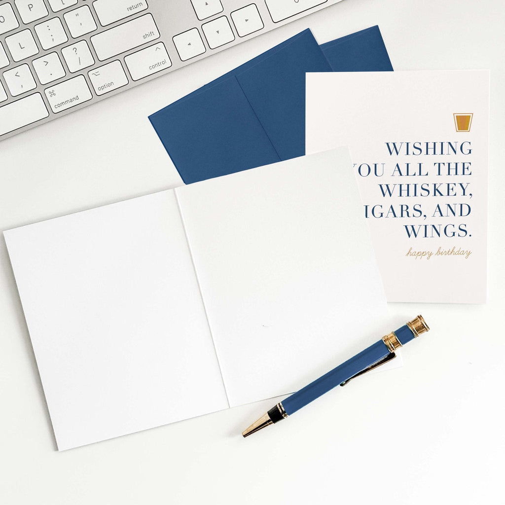 Whiskey Birthday card by Blú Rose shown open with blank interior, blue envelope and pen on a white desk