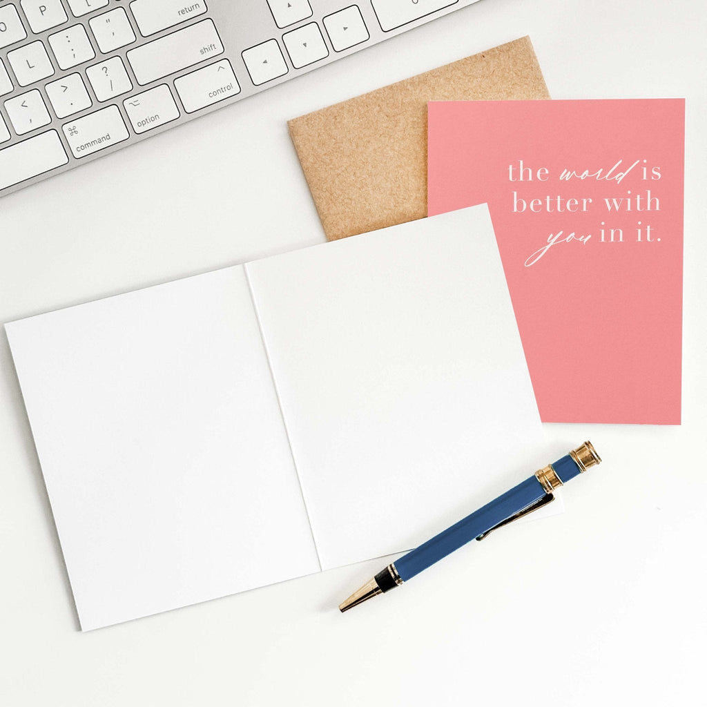 World is Better With You encouragement card by Blú Rose shown open with blank interior, kraft envelope and pen on a white desk