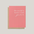 A coral pink greeting card with white script text reading 'the world is better with you in it.' shown with a kraft brown envelope on a light gray background.
