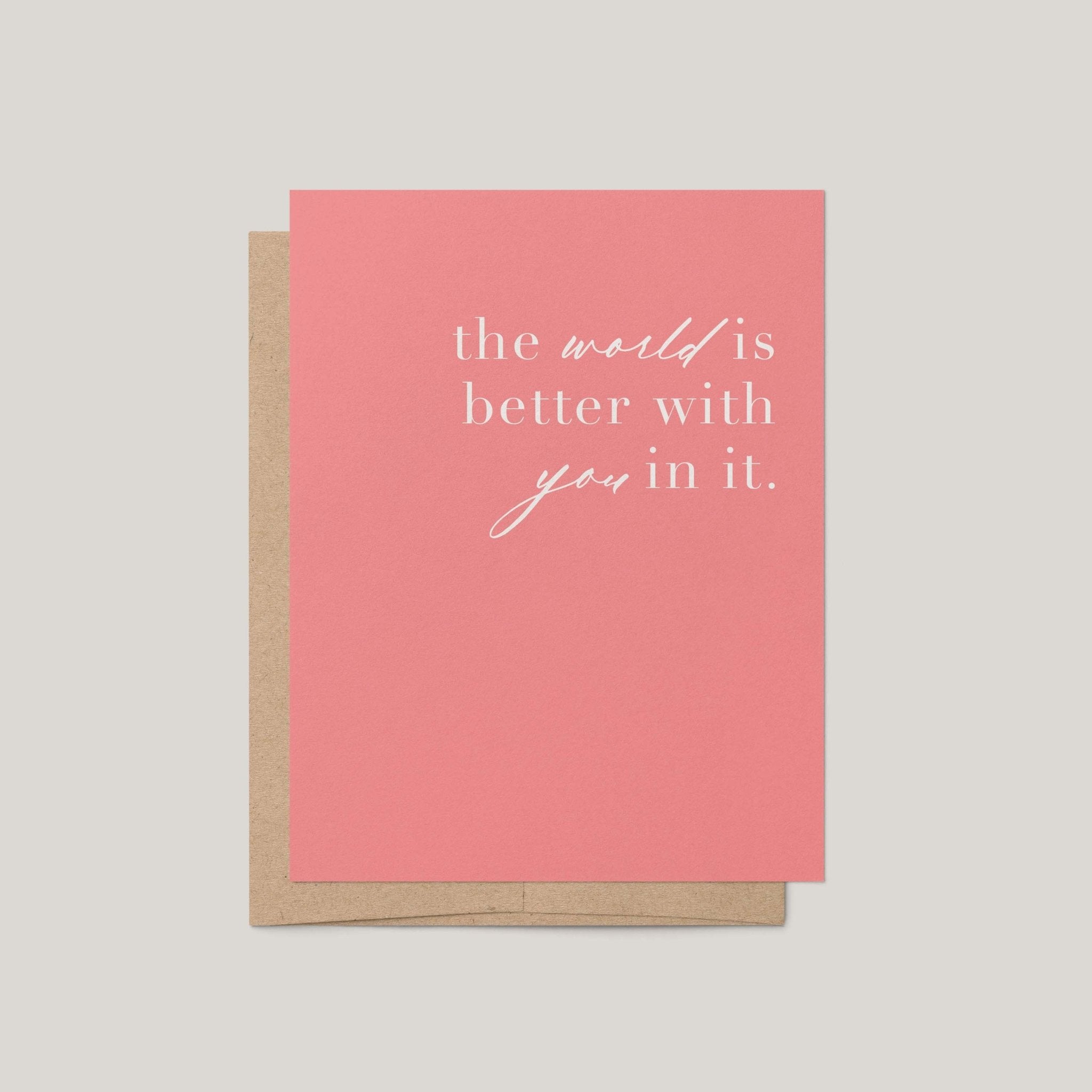 A coral pink greeting card with white script text reading 'the world is better with you in it.' shown with a kraft brown envelope on a light gray background.
