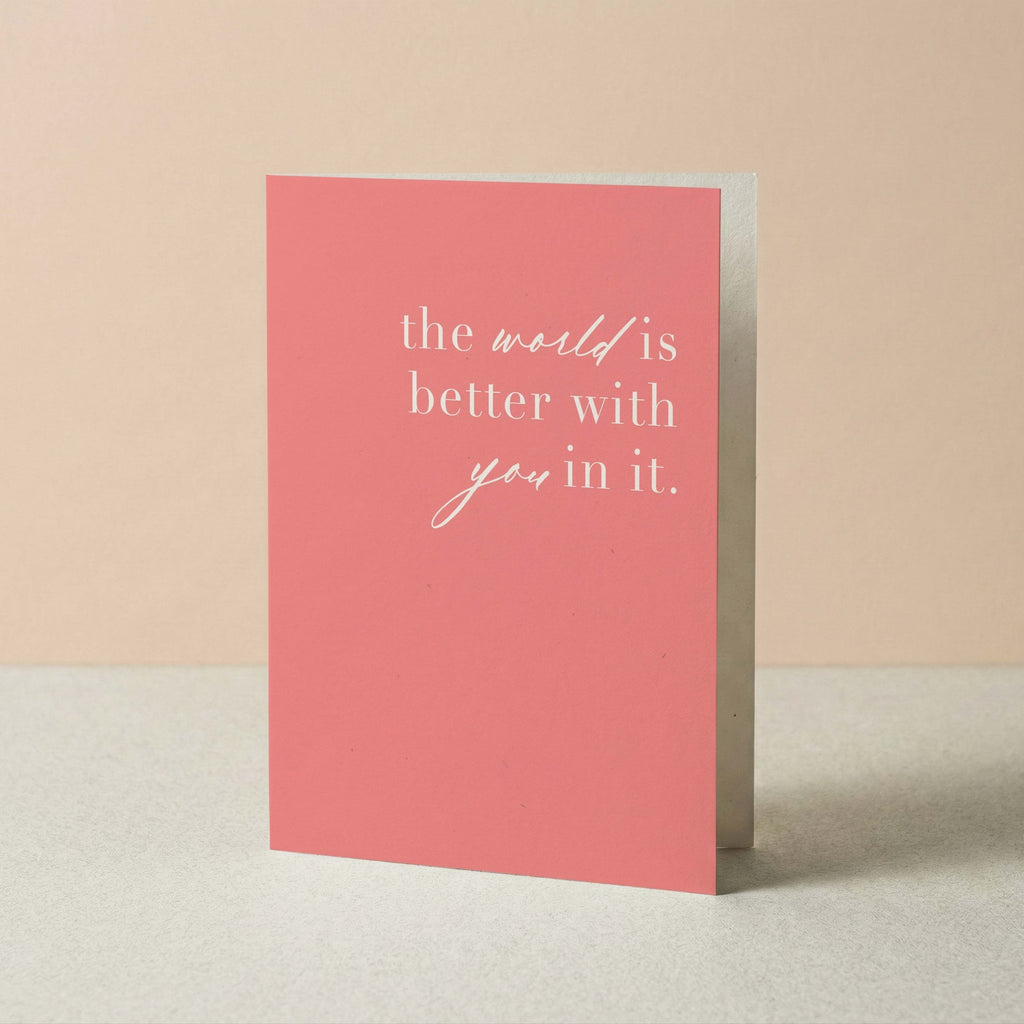 A pink greeting card with white text reading 'the world is better with you in it.' standing upright on a light surface against a soft beige background.