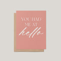 A pink greeting card with white text reading 'YOU HAD ME AT hello' layered over a kraft brown envelope, shown on a light gray background.