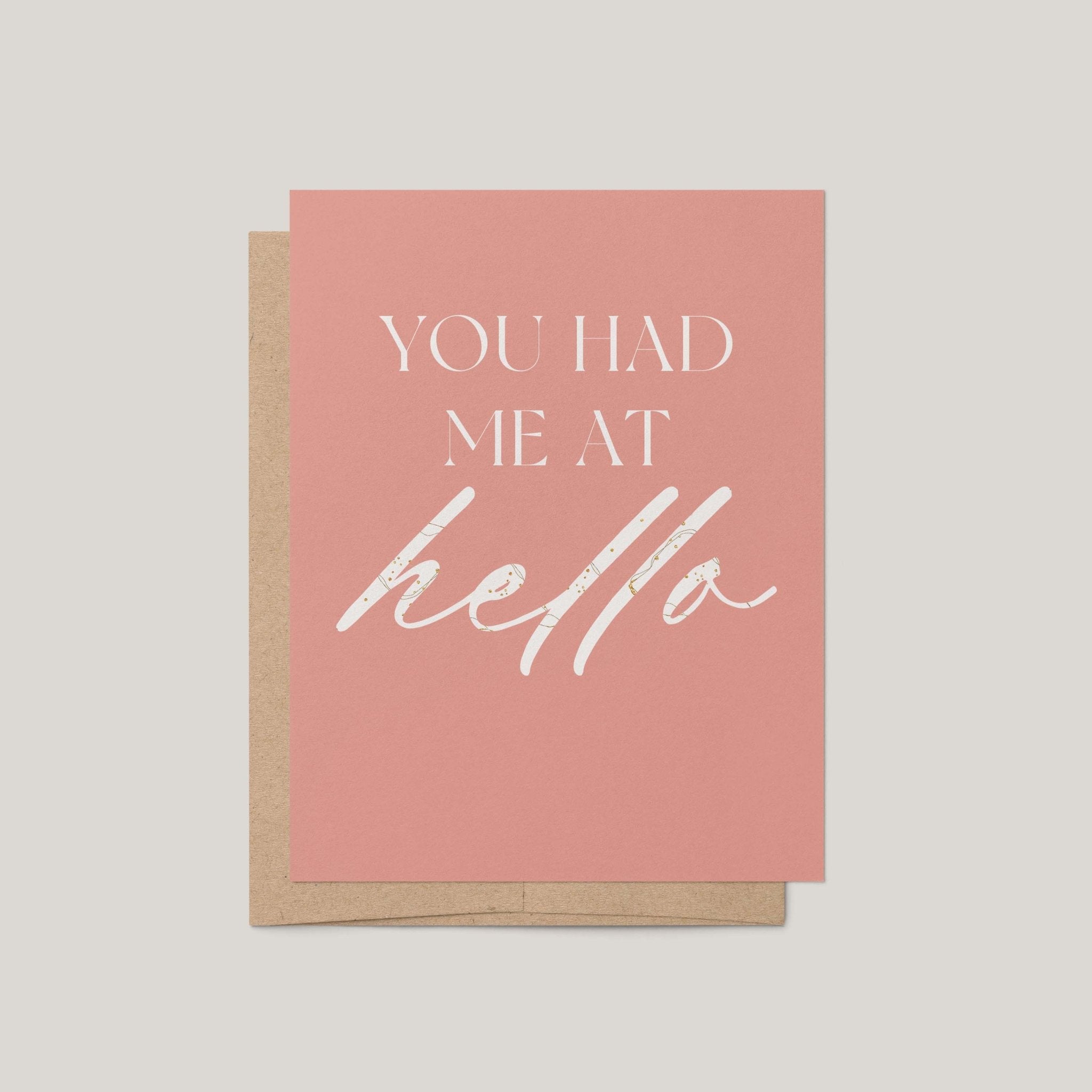 A pink greeting card with white text reading 'YOU HAD ME AT hello' layered over a kraft brown envelope, shown on a light gray background.