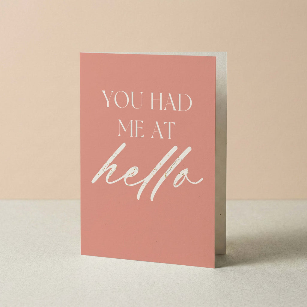 A pink greeting card with white text reading 'YOU HAD ME AT hello' standing upright on a light surface against a soft beige background.