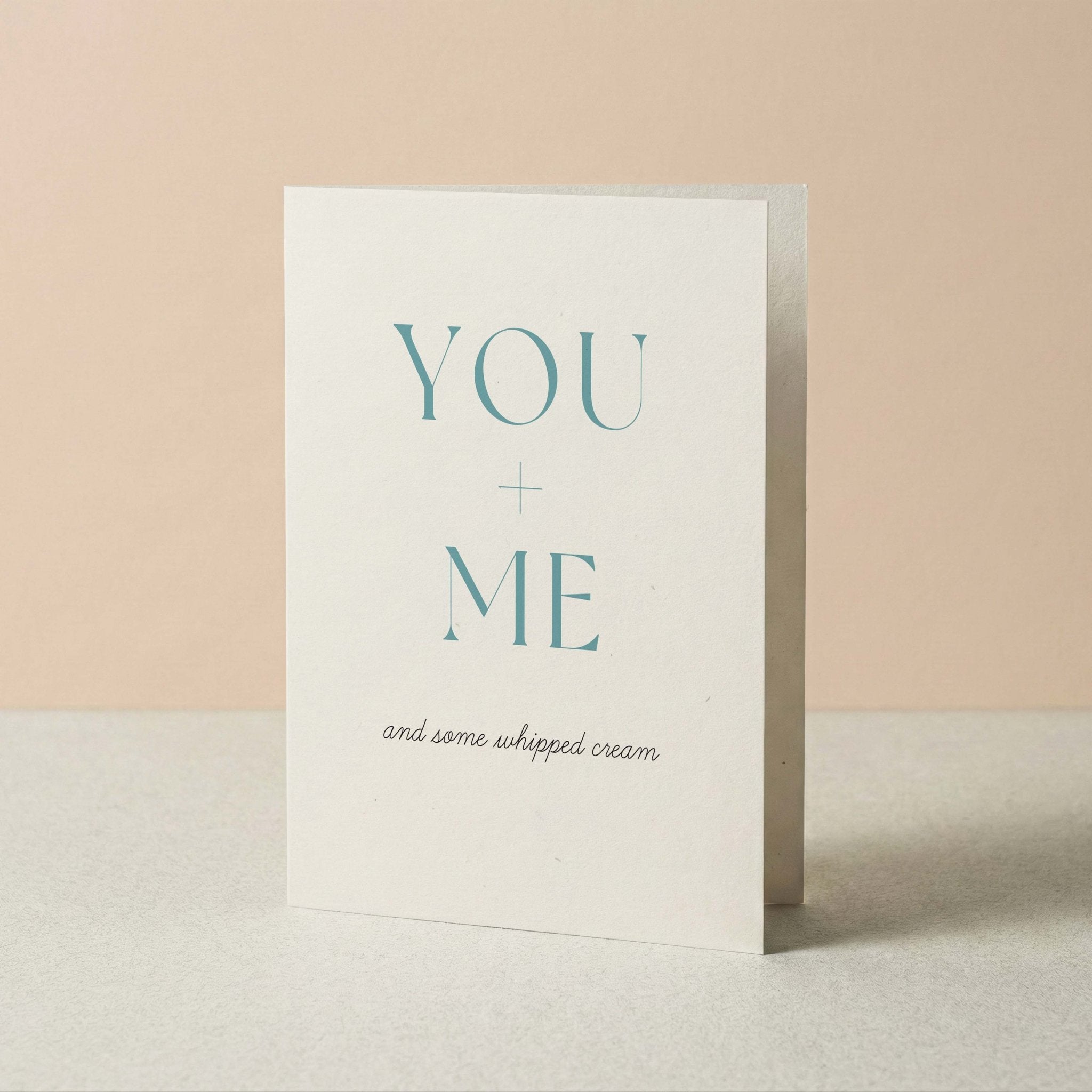 A white greeting card standing upright on a light surface against a soft peach background. The card reads 'YOU + ME and some whipped cream' in teal and black text.