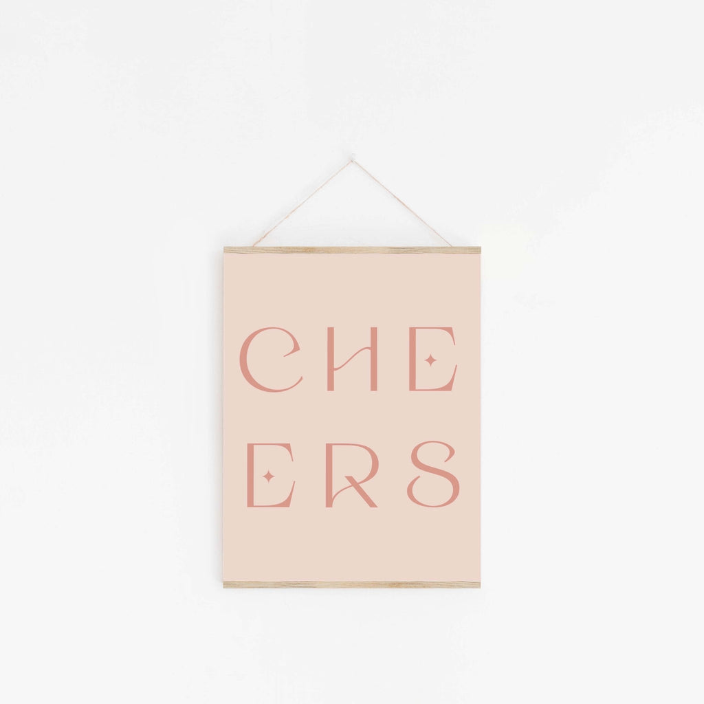 Pastel pink “CHEERS” typography art print hanging on white wall.