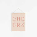 Pastel pink “CHEERS” typography art print hanging on white wall.