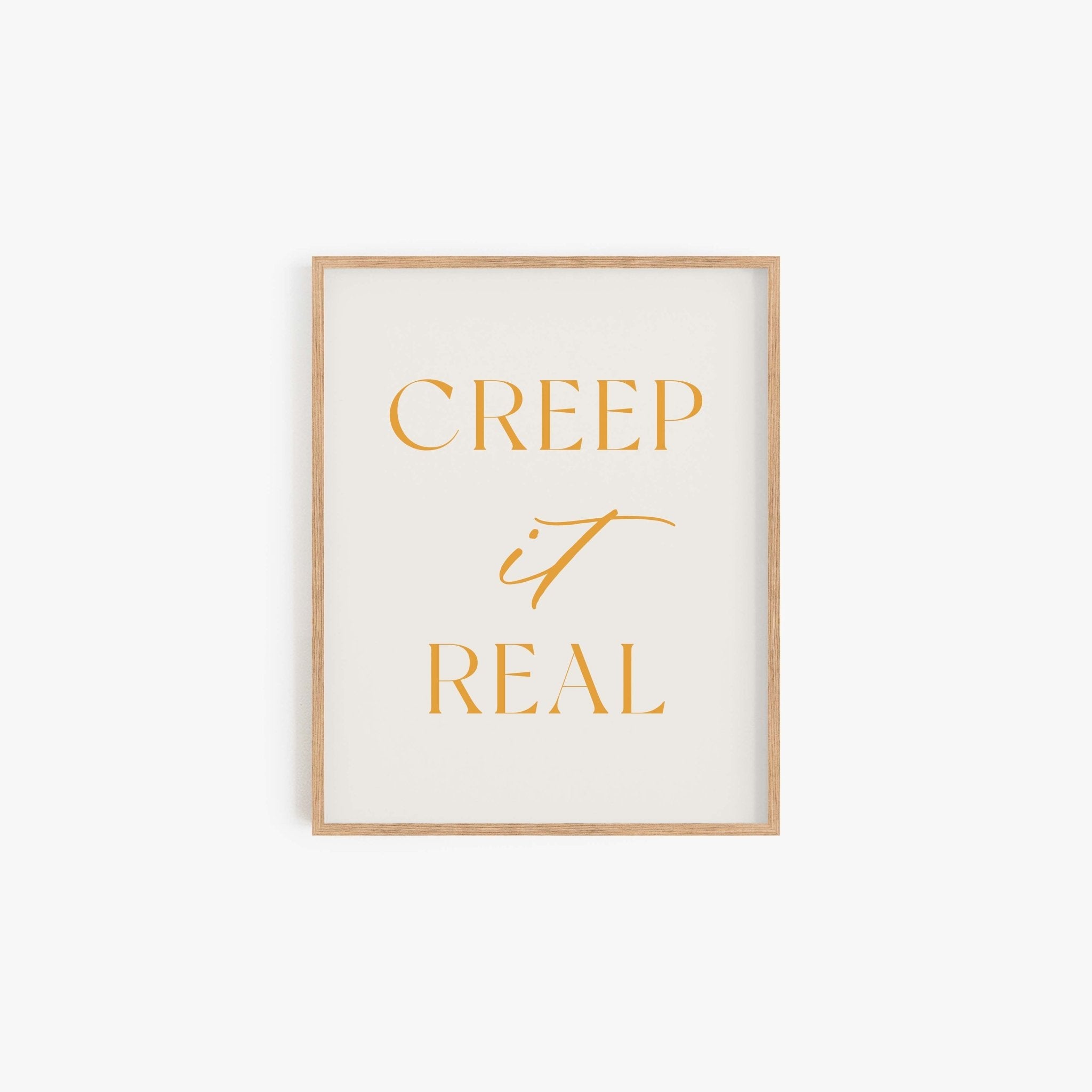 Minimalist art print on a white background that reads “Creep it Real” in marigold lettering, framed in light wood.